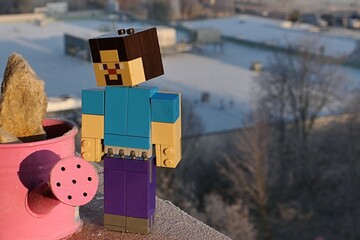 Obraz premium LEGO Minecraft large figure of smiling Steve adoring frosted pink watering can, while standing on balcony cornice, winter building in background. Morning sunshine. 