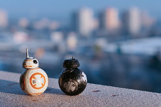 Models of Star Wars droids BB8 and imperial BB9E standing against each other on frosted winter balcony cornice, winter cityscape in background. Morning daylight sunshine. 