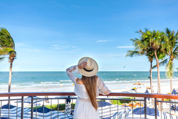 Woman in white dress with straw hat, observing sea view from hotel balcony with palms and beach view. Resort leisure and relaxation.