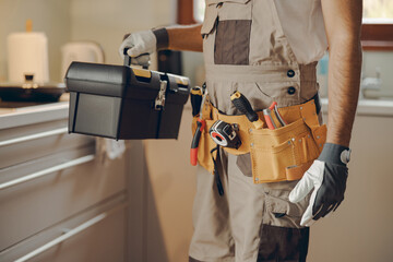 Close up of repairman in uniform standing on home kitchen and holding his tool bag