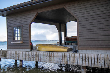 View of a large wooden open-sided boathouse, with an upside down skiff, water and clouds in...