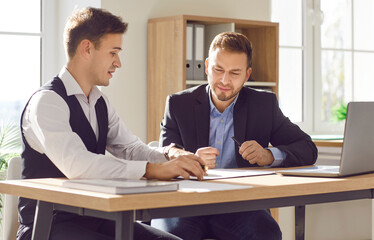 Two confident business men at work in office. Young company employees sitting at the desk on workplace having discussion, making a deal or reaching agreement and going to sign a contract.