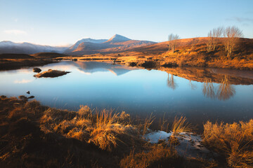 Gold light cast across a calm, tarn reflection and moorland at Rannoch Moor in the mountain landscape of Glencoe in the Scottish Highlands, Scotland, UK. - obrazy, fototapety, plakaty