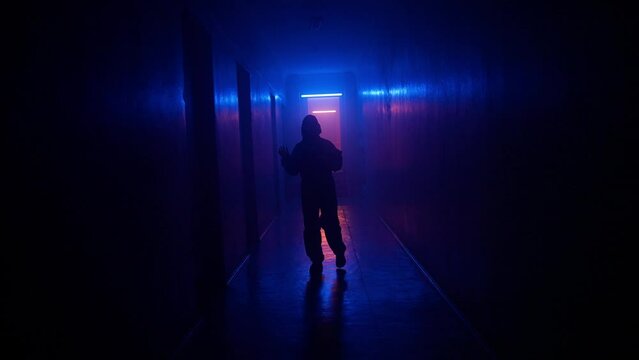 Portrait of silhouette in the hallway neon light. Person in chemical protection suit walks cautiously in the dark corridor, looking around facing camera.