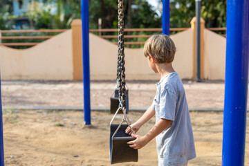 Thoughtful preschooler boy, stands near swing, as if deciding whether to try to swing on them.