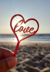 Person holding in fingers hand stick in shape red heart and word Love on background sea and sea...