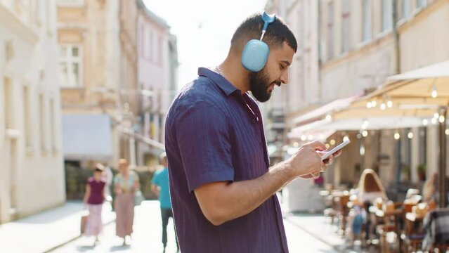 Happy relaxed overjoyed indian man in wireless headphones choosing, listening favorite energetic disco music in smartphone dancing outdoors. Guy tourist walking passes by urban sunshine city street
