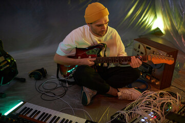Medium long shot of male musician surrounded by musical equipment checking electric guitar strings before recording while sitting on studio floor