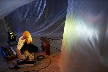 Wide angle shot of male musician changing settings of sound mixer while sitting on floor of improvised studio covered with plastic