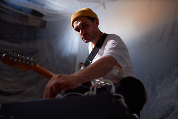 Low angle view of Caucasian male musician holding electric guitar on strap for record session while adjusting musical equipment settings