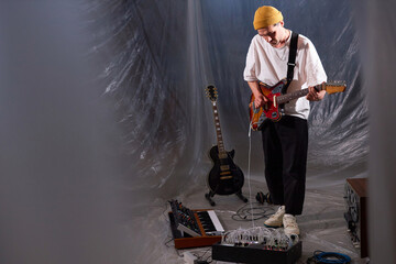 Full shot of male musician standing in makeshift studio with plastic cover while playing electric guitar connected to sound mixer for audio samples recording