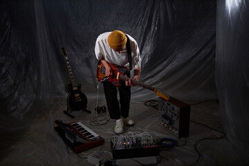 Wide shot of unrecognizable male musician standing half-bent playing electric guitar in dim light portable recording studio covered with transparent plastic