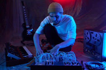 Medium full shot of male composer in blue neon light adjusting portable sound mixer for recording session while sitting on floor of improvised studio