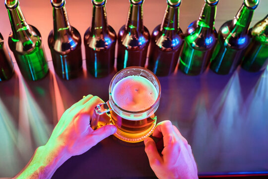 Uma mão segurando uma caneca com chope e muitas garrafas ao redor sobre o balcão de um pub.