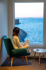 Woman in pajamas reading in a window facing the sea at sunset