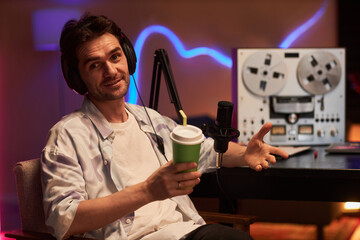 Portrait of brunette male interviewer with studio headphones looking at camera talking and gesturing while sitting in podcast room