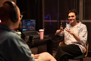 Medium long shot of beaming adult male podcaster having fun talk with unrecognizable guest while sitting legs crossed in studio room