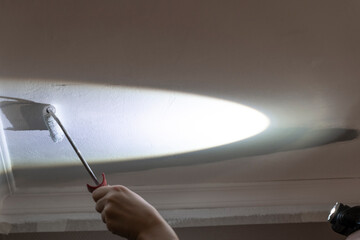 Ceiling Brilliance: Woman Paints with Precision, Using Roller and Headlight for Enhanced Visibility.