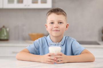 Cute boy with glass of milk at white table in kitchen