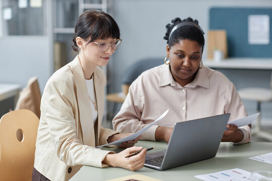 Medium shot of two smiling diverse female coworkers holding papers looking at laptop while sitting at desk in office