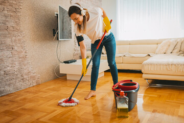 The young woman is wiping the parquet floor in the living room and wearing yellow glove