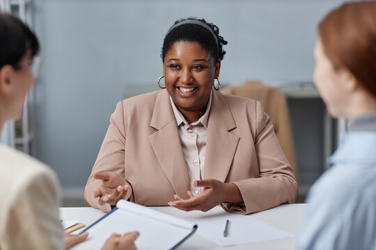 Medium shot of laughing African American female candidate wearing pastel suit gesturing and telling about herself during job interview with unrecognizable HR agents in office