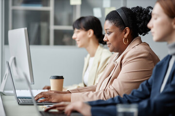 Side view of female company employees working on computers in office, focus on smiling adult African American business woman dressed in pastel suit looking at laptop screen - Powered by Adobe