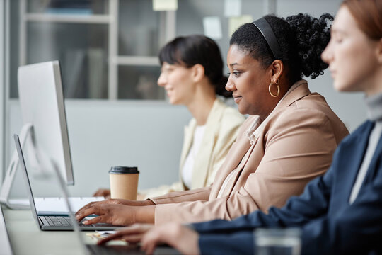 Side View Of Female Company Employees Working On Computers In Office, Focus On Smiling Adult African American Business Woman Dressed In Pastel Suit Looking At Laptop Screen