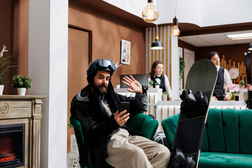 Caucasian male guest seated on sofa surrounded by snowboarding gear, having a video call on his digital phone. Tourist in snow clothing on videoconference with mobile device in ski resort lounge area. - obrazy, fototapety, plakaty