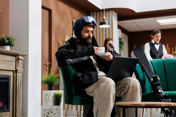 In luxury lounge area, man dressed in winter jacket enjoys coffee while using laptop to explore...