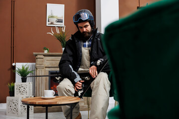 Caucasian male skier in winter clothing fine-tuning his snow equipment in ski resort lounge area, ready for snowy adventure. Youthful man seated near fireplace in hotel lobby is securing skiing skis. - obrazy, fototapety, plakaty