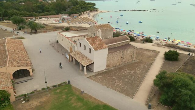 Aerial orbit around romanesque Church of Sant'Efisio (Chiesa di Sant'Efisio) in Nora, Pula, Sardinia, Italy. Beautiful beach behind, with colorful umbrellas, boats and people swimming.