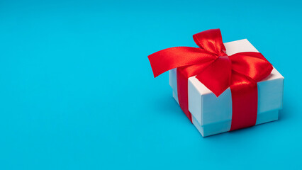 Close up cropped shot of a gift box wrapped in white paper and decorated with red satin ribbon isolated over blue background with a copy space
