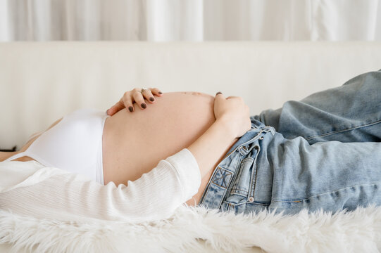 Cropped color photo side view of pregnant woman in unbuttoned jeans laying on white couch with hands on her bare belly 