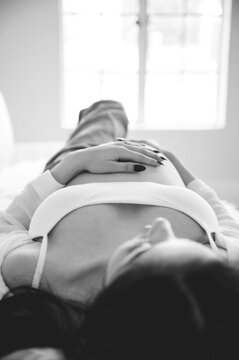 Cropped black and white photo of pregnant woman in unbuttoned blue jeans laying on white couch with hands on her bare pregnant belly aesthetic