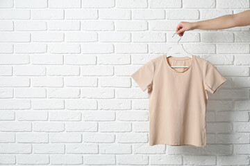 Woman holding hanger with beige T-shirt near white brick wall, closeup. Space for text