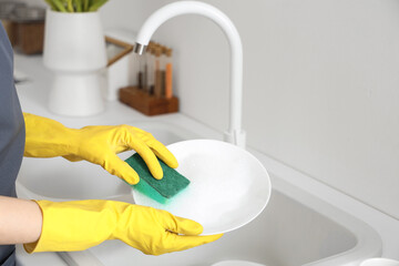 Woman in yellow rubber gloves washing plate with sponge in light kitchen