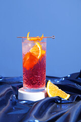 Glass of cold Negroni cocktail with orange on shiny fabric against blue background