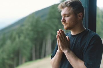 A man is praying near the beautiful panoramic window against the background of the forest. Christian man. Concept for faith, spirituality and religion.