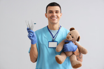 Young male dentist with professional tools and teddy bear on grey background. Dental care concept