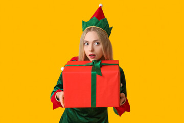 Young woman in elf costume with Christmas gift box on yellow background
