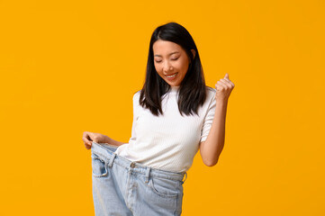 Beautiful young happy Asian woman in loose jeans on yellow background