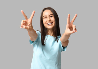 Beautiful young woman showing victory gesture on grey background