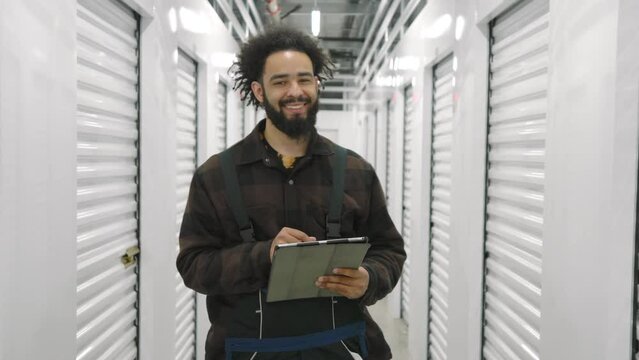 Bearded smiling dark-skinned man, self storage worker in checkered T-shirt, work overall holding tablet in a typical climate controlled storage facility. High quality 4k footage
