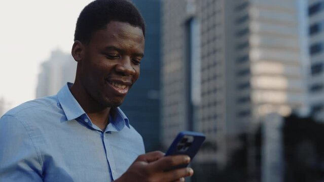 Happy African American Manager Standing Outside and Browsing Internet on his Phone. Smiling Businessperson Outdoor Watching the Video in Mobile. People and Technology Concept