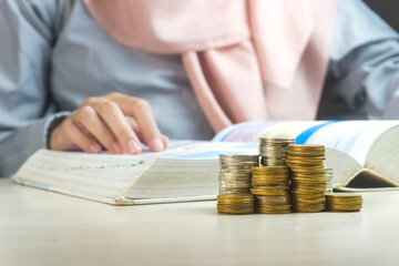 muslim businesswoman reading a book with heap of coins money in the front symbol for learning...