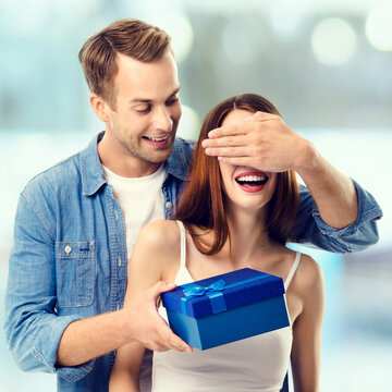 Consumer credit, celebrating, sales, rebates concept - happy amased couple with blue gift box. Man and woman standing over blurred modern interior or mall.