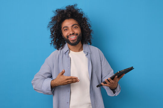 Young joy ethnic Arabian man brunette points hand at tablet computer urging you to use gadget to download educational applications or surf social networks stands on plain blue background.