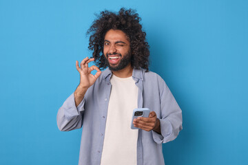 Young successful excited Arabian man holds smartphone and shows approving gesture looking at screen and winking as sign of support for new version of mobile phone stands posing in blue studio. - obrazy, fototapety, plakaty