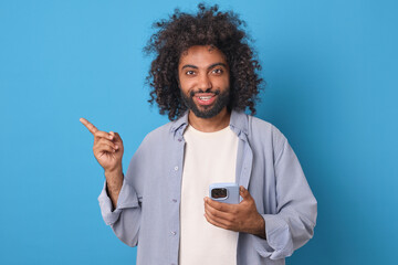 Young happy handsome Arabian man with mobile phone in hands smiles sincerely and points finger to side offering to download online store application for smartphone users stands in blue studio.